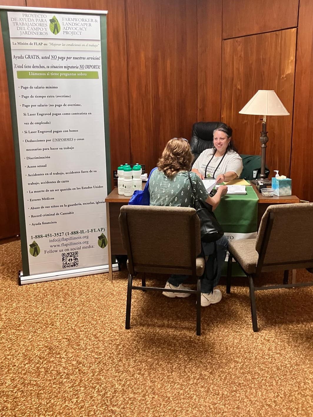 A FLAP staff member meets one-on-one with a woman across a desk, beside a Spanish-language banner listing the workplace problems FLAP can help with.