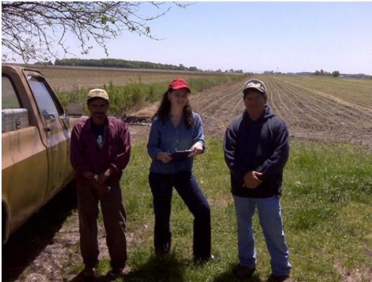 A FLAP staff member stands with two farmworkers beside a pickup truck at the edge of a field.