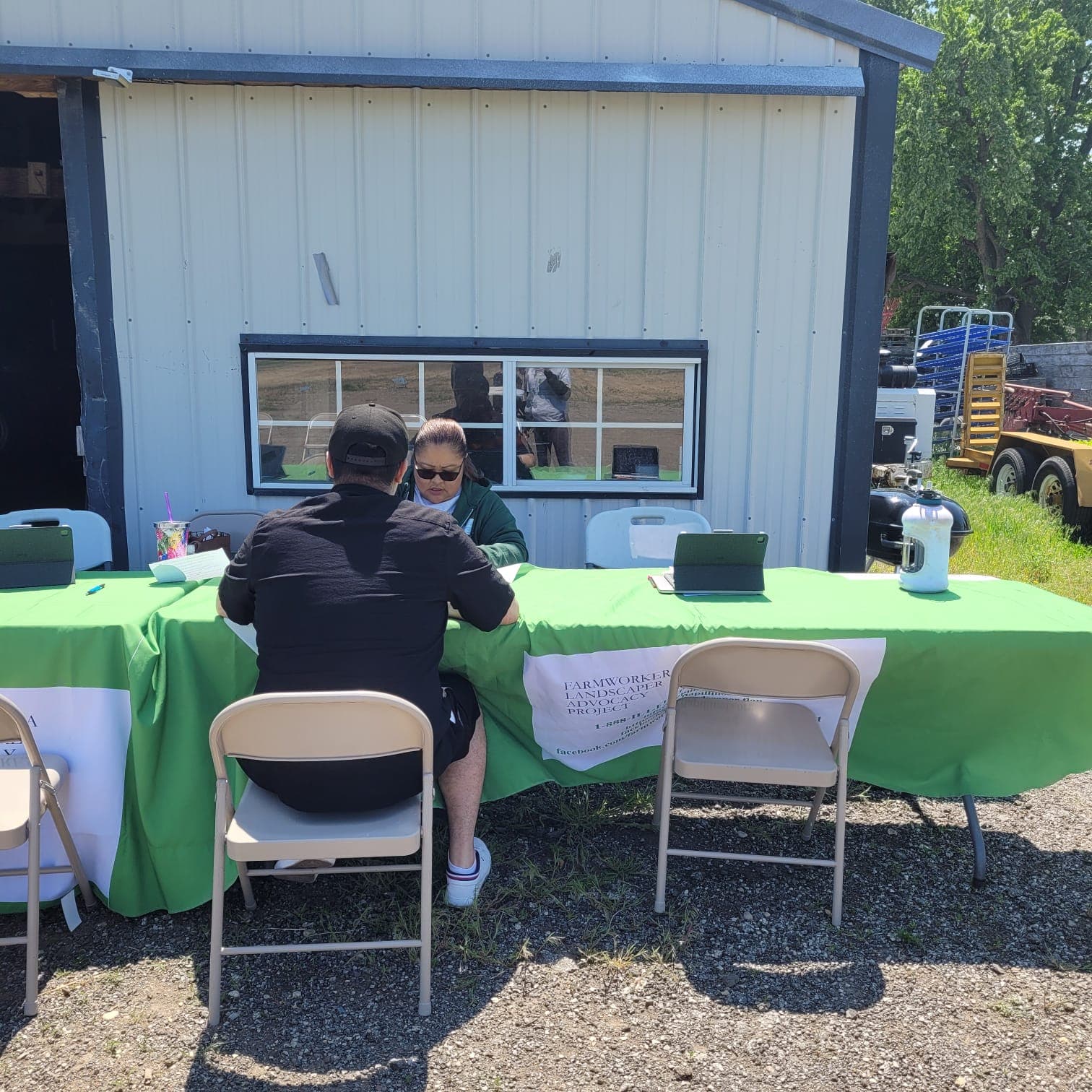 A FLAP staff member meets with a worker at an outdoor intake table set up beside a farm shed.