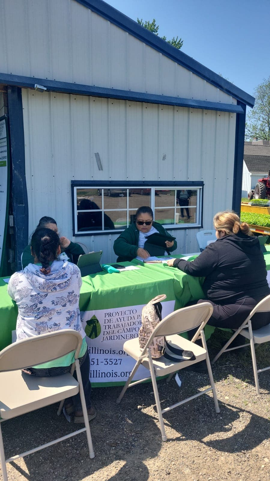 FLAP staff meet with farmworkers at a green-draped intake table outside a farm building.