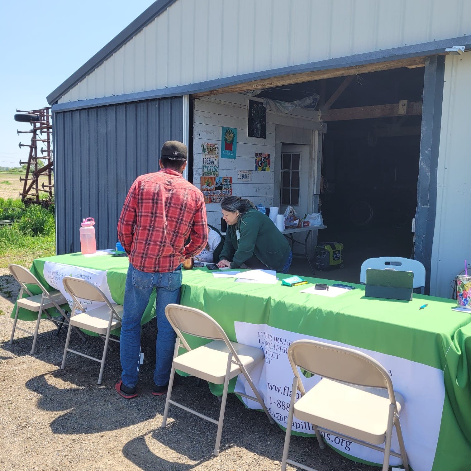 A farmworker signs in with a FLAP staff member at an outdoor table beside a farm shed.