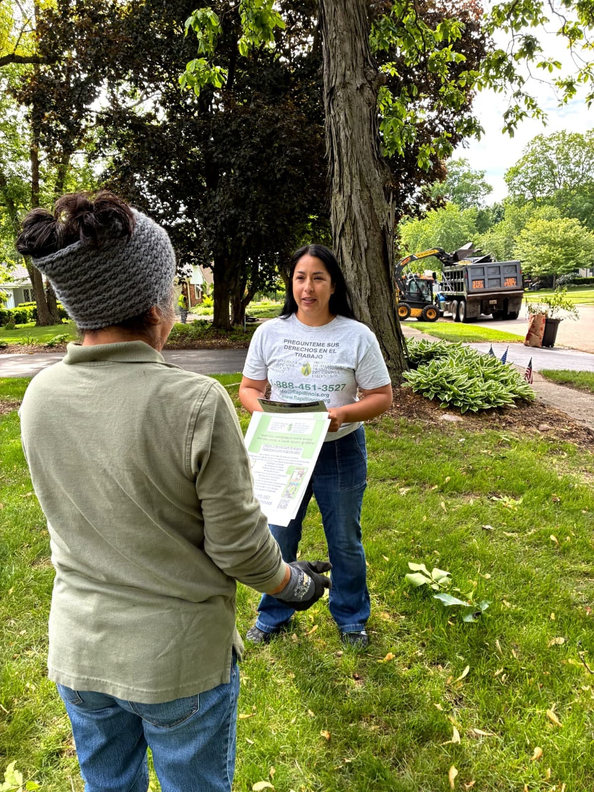 A FLAP outreach worker hands a flyer to a landscaper working on a tree-lined residential street.