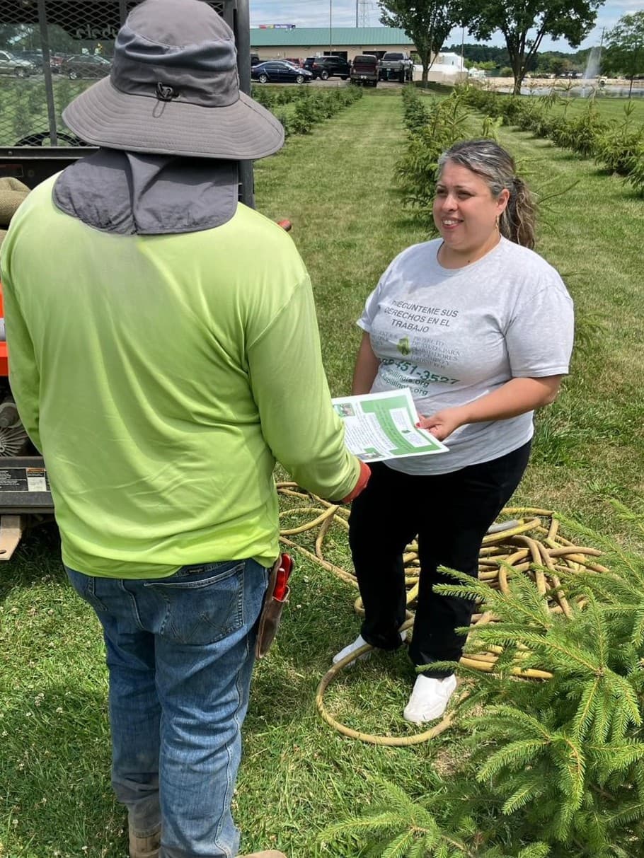 A FLAP outreach worker gives a know-your-rights flyer to a nursery worker among rows of young trees.