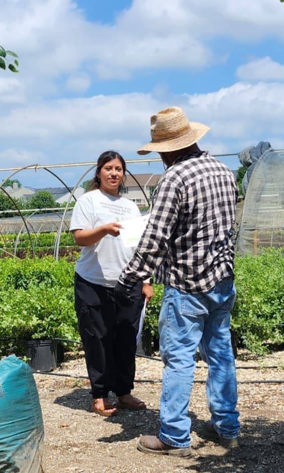 A FLAP outreach worker hands a flyer to a farmworker beside the greenhouse rows of a plant nursery.