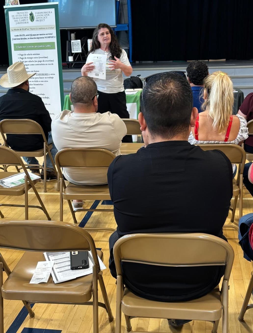 A FLAP attorney presents to workers seated in a school gymnasium, beside FLAP's Spanish-language banner.