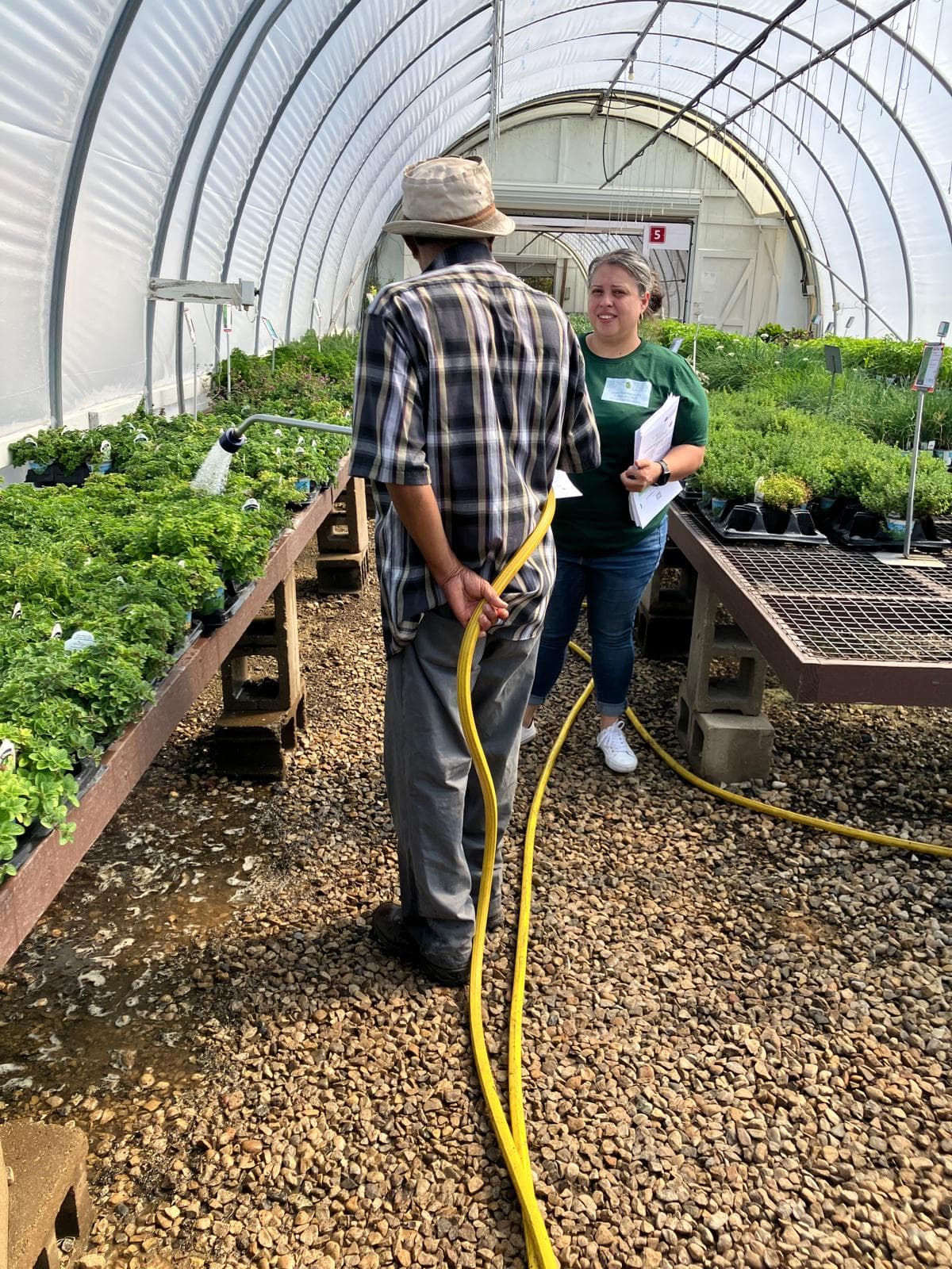 A FLAP outreach worker speaks with a nursery worker watering plants inside a greenhouse.